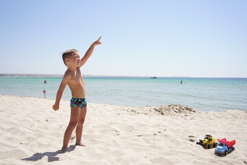 Happy baby playing with cars on the beach, on the horizon azure sea and blue sky. A child stands on the beach and points his finger at the sky, he saw the plane.