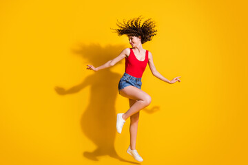 Full size photo of lovely crazy young lady closed eyes jump raise hands festival dance shake head wear jeans shorts red singlet uncovered shoulders isolated vibrant yellow color background
