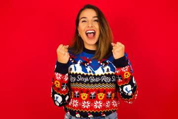 Young beautiful Caucasian woman wearing Christmas sweater against red  rejoicing success and victory clenching his fists with joy being happy to achieve her aim and goals. Positive emotions, feelings.