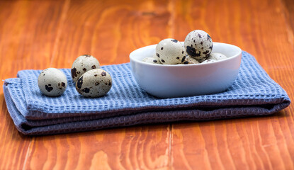 Quail eggs in white ceramic bowl on blue kitchen cloth. Top view. Detailed closeup of spotted quail eggs, rustic wooden background