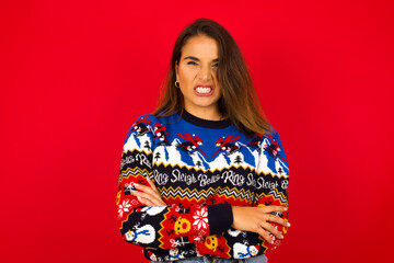 Mad crazy Young beautiful Caucasian woman wearing Christmas sweater against red wall,  clenches teeth angrily, being annoyed with coming noise. Negative feeling concept.