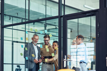 Brainstorming. Group of multiracial creative coworkers standing in the modern office and looking at colorful sticky notes on a glass wall, discussing new project