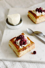 Puff pastry squares with ricotta and cherry sauce on white marble tray. Baked small square cakes topped with cheese and berry sauce