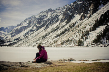 Beautiful young woman sitting next to stunning winter landscape. Concept of travel and adventure at the frozen Gaube Lake (Lac de Gaube) in the French Pyrenees, Cauterets, France.