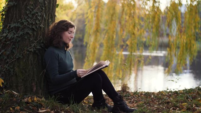 Beautiful young woman sitting under tree reading a book during sunset. Attractive nature-loving millennial girl relaxing and studying outdoors in a park with a weeping willow and lake. Slow-motion.