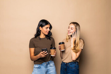 Portrait of two smiling women using cellphones while drinking coffee takeaway isolated over beige background