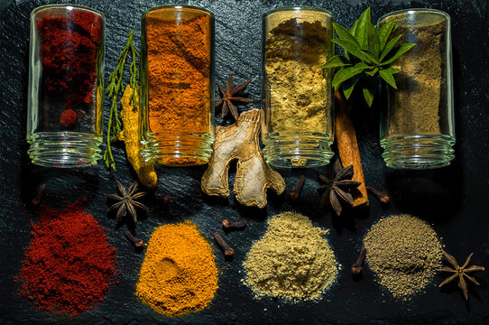 Variety of Spices Used in Cooking, Turmeric, Ginger, Paprika, Pepper, presented with Clear Glass Jars on Black Stone Texture Background, Surrounded by Ginger, Turmeric Root, Star Anise and Clove.