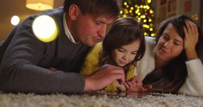 Close up of happy Caucasian family with little daughter laying on floor in cozy decorated room typing and tapping on tablet together on Christmas Eve together. Family time. New Year