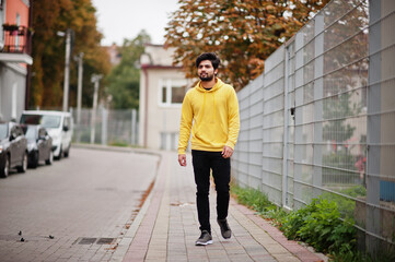 Urban young hipster indian man in a fashionable yellow sweatshirt. Cool south asian guy wear hoodie walking on fall street.