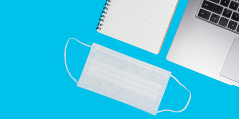 Flat lay composition with laptop, medical face mask, notepad isolated on blue background top view with copy space. Concept of safety work in coronavirus pandemic