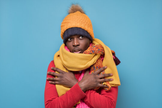 Funny African American Mature Man In Yellow Scarf And Hat Freezing In Warm Clothing. Studio Shoot On Blue Wall.