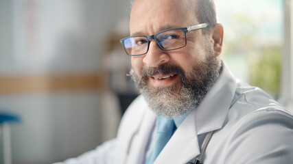 Fototapeta premium Latin Doctor's Office: Experienced Physician Wearing Glasses Sitting at His Desk Working, Turns, Looking at Camera, Smiling Charmingly. Health Care Specialist Checking Test Results. Close-up Portrait