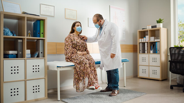 Latin Doctor's Consultation Office: Physician Uses Stethoscope to Listen to Heartbeat and Lungs of the Female Patient. Medical Health Care Professionals Diagnosing Disease. Both Wearing Face Masks