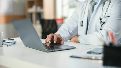 In a Doctor's Consultation Office: Experienced Physician Working on a Laptop, Prescribing Medicine, Analysing Tests Results. Close-up Shot of a Hand and Laptop.