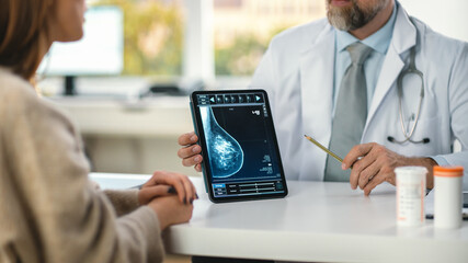In a Doctor's Consultation Room: Friendly Professional Doctor Uses Digital Tablet Computer to Show Breast Scan to a Female Patient and Explains Importance of Breast Cancer Prevention. Focus on Tablet - obrazy, fototapety, plakaty