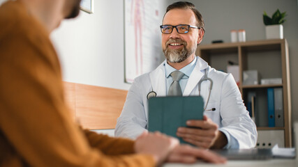 Obraz premium In a Doctor's Consultation Office: Professional Physician Smiles Charmingly Talks to the Patient, Uses Digital Tablet Computer to Ask Questions about Symptoms. He is Checking Patient's Treatment Plan