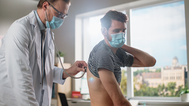 Doctor Consultation Office: Physician Listens To Patient's Lungs And Heart From The Back. Both Wear Face Masks. Medical Health Care Specialist Works With Breathing Problems, Prescribes Treatment