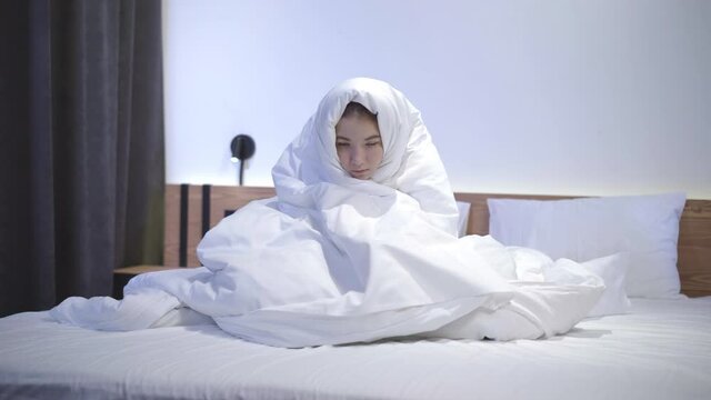 Wide shot of ill teenage Caucasian girl freezing in bedroom. Portrait of sad beautiful teenager wrapping in blanket or duvet. Fever and symptoms of Covid-19 viral disease. Coronavirus pandemic.