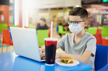 Fototapeta premium Teenager wearing glasses and a protective mask uses a laptop in a restaurant before lunch