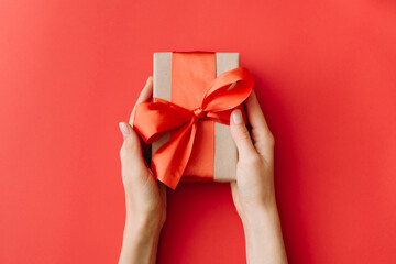Woman hands holding present box with red bow on red background. Flat lay, top view.
