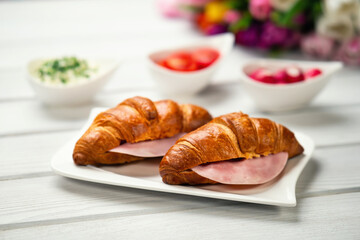 Fresh croissant sandwich with ham, tomatoes and radish on white table. Healthy romantic breakfast for two with bouquet in the background