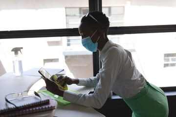African american woman wearing face mask cleaning her digital tablet with cloth at modern office