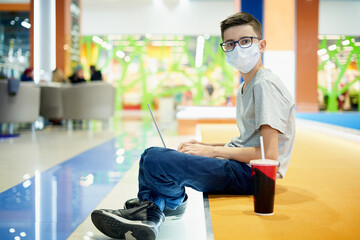 Portrait of a teenage boy in a protective mask with a laptop in a shopping mall