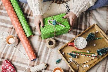 Young woman sits on plaid in cozy knitted woolen white sweater, socks and wraps Christmas gift in polka dot wrapping paper. Wooden tray with mug of cocoa with marshmallows, toy tree, candle, straws.