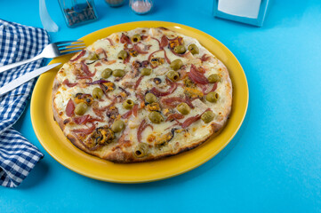 Classic pizza with mussels, meat, green olives, cheese. yellow plate on a blue table. Cafe menu. Fork, knife and place for text. Food with seafood and olive. Copy space.