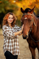 Obraz premium a young beautiful brunette woman in a plaid shirt holds a brown horse by the bridle