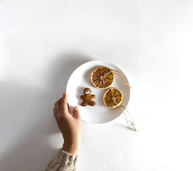 Woman's hand in a knitted sweater holds a plate of Christmas sweets. Cookies in the form of a man in a mask and dried orange slices with cinnamon. On a white background