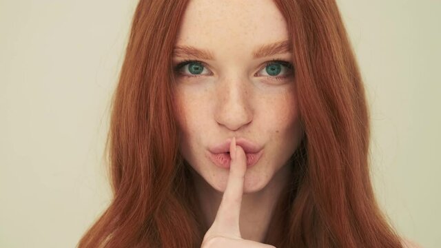A close-up view of a positive woman is doing secret gesture standing isolated over beige background in studio