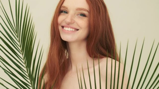A smiling woman is posing to the camera while looking through palm leaves standing isolated over beige background in studio