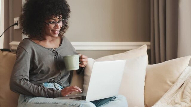 Young cheery african woman sitting indoors at home and using laptop computer while drinking coffee