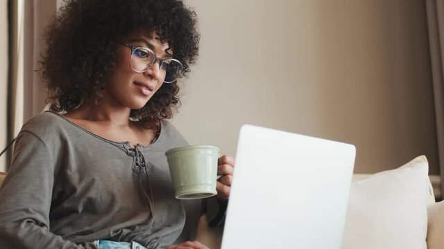 Young happy african woman sitting indoors at home and using laptop computer while drinking coffee