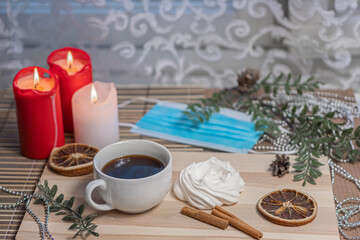 Medical mask, Christmas candles, coffee mug on a wooden table, air meringue, dried orange, cinnamon and accessories for winter holidays. New Year