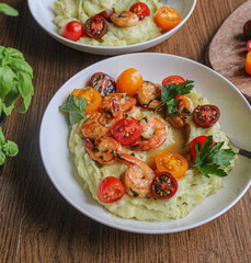 Glazed shrimp with garlic and honey sauce over creamy mashed potatoes and topped with colourful cherry tomatoes and coriander leaves. Beautiful spring meal plated on wooden table. 