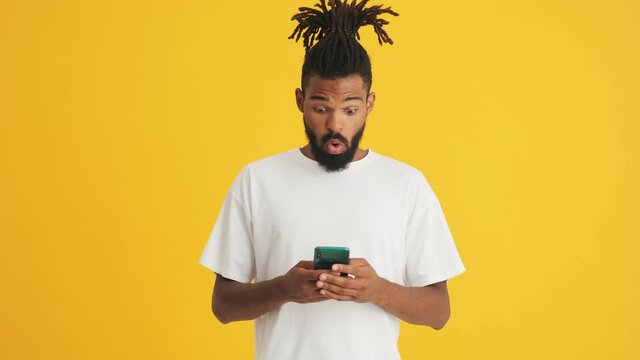 Young handsome shocked african man using mobile phone isolated over yellow background