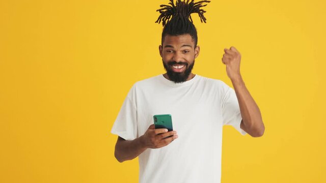 Young handsome african man using mobile phone and showing winner gesture isolated over yellow background