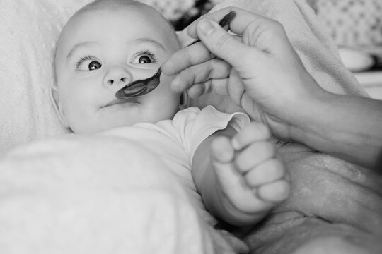 Mother feeding her infant son with spoon. Cute little 6 month old baby boy eating with funny grimace. Black and white photo.