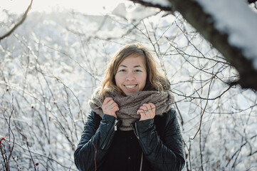 Beautiful happy young girl in winter clothes with a scarf on a background of trees covered with snow in the park. Portrait of happy woman.