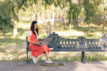 Chica trabajando en el parque con su computadora portátil, sentada en una banca con arboles a los...