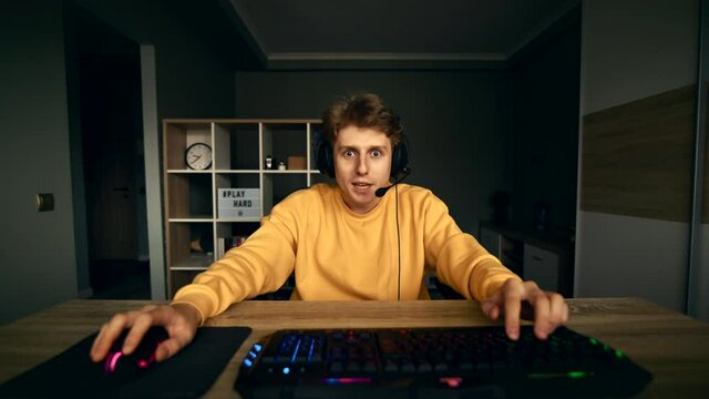 Joyful gamer in a headset sits at home at the computer and rejoices in winning a computer game. Winning gamer rejoices and communicates with friends on the microphone