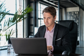 Young businessman wearing black suit, sitting to table with laptop. Office manager working in...