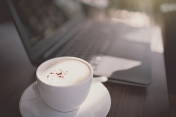 White coffee cup on wooden table with blurred computer notebook background. Drama tone.