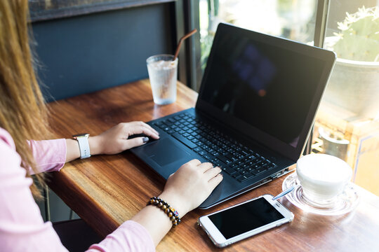 Mockup image of an Asian woman sitting on table, using and typing on laptop with blank black desktop screen keyboard and smart phone and coffee cup.
