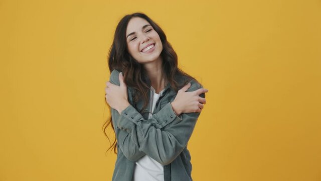 A smiling woman is hugging herself standing isolated over yellow background