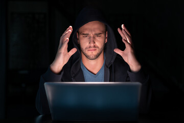 Stressed hacker in black hoodie with laptop hands in the sky, in dark room, man with a hood, programming late at night. Frustrated man hacks a computer laptop at night. Concept of hacker