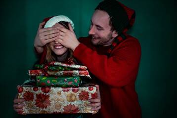 Boyfriend covering the eyes of the girlfriend while they wear christmas outfit. Boyfirend surprising girlfriend with a lot of gifts for christmas.