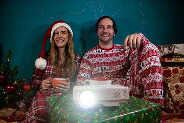 Young cheerful couple watching movie on projector with christmas pijama.